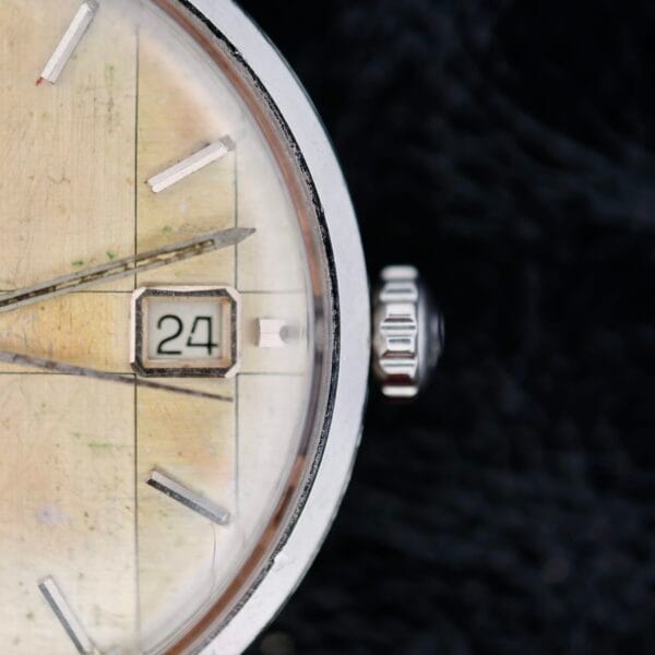 A close-up of an Omega Seamaster Deville Automatic 166.020 showcases a vintage analog watch face with a worn, off-white background, the time set at approximately 10:10 and the date reading 24. The silver case and crown provide elegance against a dark, textured backdrop.