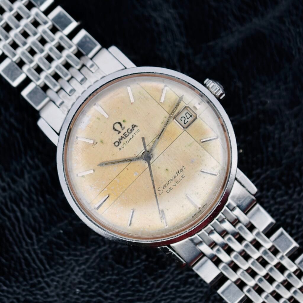 The Omega Seamaster Deville Automatic 166.020, featuring a vintage round yellow dial with silver hour markers and hands, plus a date window at 3 oclock, is showcased on a dark textured background. It includes a silver metal mesh bracelet.