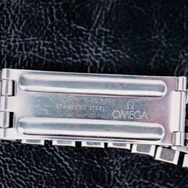 A close-up of the Omega Seamaster Deville Automatic 166.020 stainless steel watch bracelet shows an open clasp with STAINLESS STEEL engraved and the logo with OMEGA beneath, set against a textured black background.