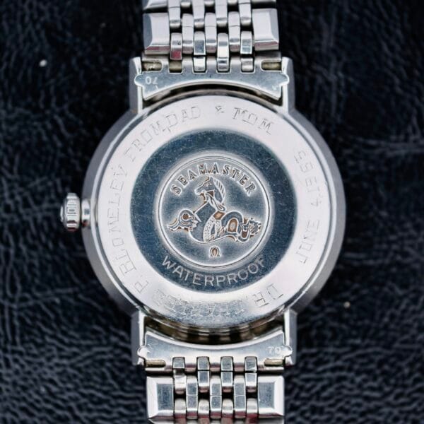 The image displays the back case of an Omega Seamaster Deville Automatic 166.020 wristwatch, featuring a stainless steel design with a seahorse emblem and SEAMASTER engraving. It includes technical details and has a metal link bracelet against a black background.