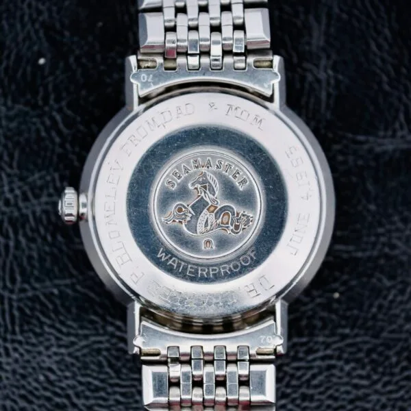 The image displays the back case of an Omega Seamaster Deville Automatic 166.020 wristwatch, featuring a stainless steel design with a seahorse emblem and SEAMASTER engraving. It includes technical details and has a metal link bracelet against a black background.
