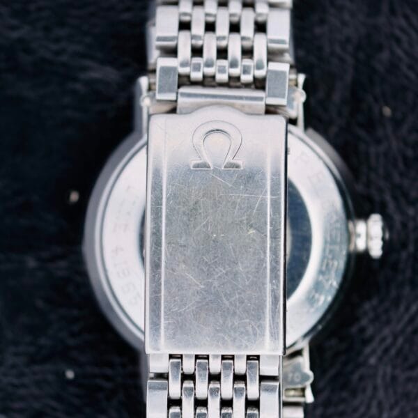 Close-up of the back of an Omega Seamaster Deville Automatic 166.020, showcasing its silver metal bracelet. The clasp displays an engraved logo like the Greek letter Omega, with visible scratches on the watch case and clasp against a dark, textured fabric background.