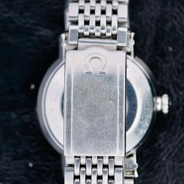 Close-up of the back of an Omega Seamaster Deville Automatic 166.020, showcasing its silver metal bracelet. The clasp displays an engraved logo like the Greek letter Omega, with visible scratches on the watch case and clasp against a dark, textured fabric background.