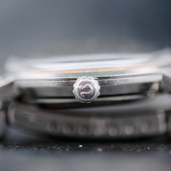 Close-up of the Omega Seamaster Deville Automatic 166.020s silver watch crown, featuring a textured edge and a small emblem. The focus is on the crown, with the rest of the watch elegantly blurred in the background, highlighting its metallic finish.