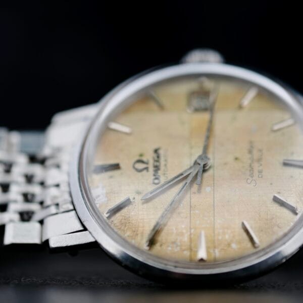 Close-up of a vintage Omega Seamaster Deville Automatic 166.020 wristwatch, showcasing its tarnished yellowed dial with silver hour markers and hands. The timepiece is attached to a metallic bracelet and presented against a dark background.