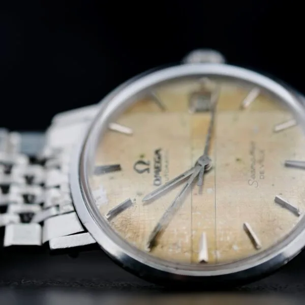 Close-up of a vintage Omega Seamaster Deville Automatic 166.020 wristwatch, showcasing its tarnished yellowed dial with silver hour markers and hands. The timepiece is attached to a metallic bracelet and presented against a dark background.