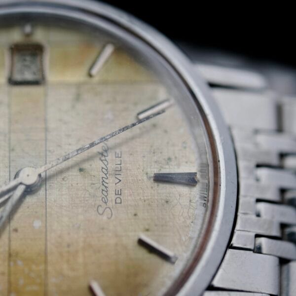Close-up of a vintage Omega Seamaster Deville Automatic 166.020 wristwatch with a yellowed face reading approximately 10:11. The watch includes a silver metal band, showing visible signs of wear and aging.