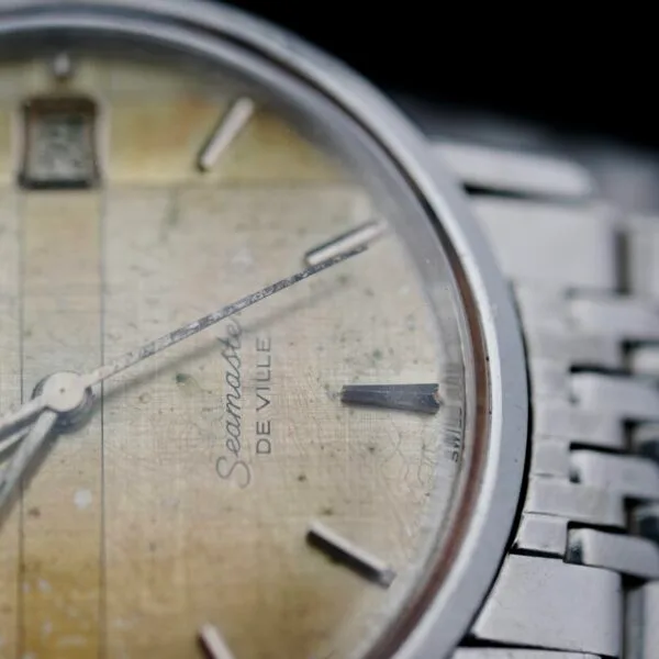 Close-up of a vintage Omega Seamaster Deville Automatic 166.020 wristwatch with a yellowed face reading approximately 10:11. The watch includes a silver metal band, showing visible signs of wear and aging.