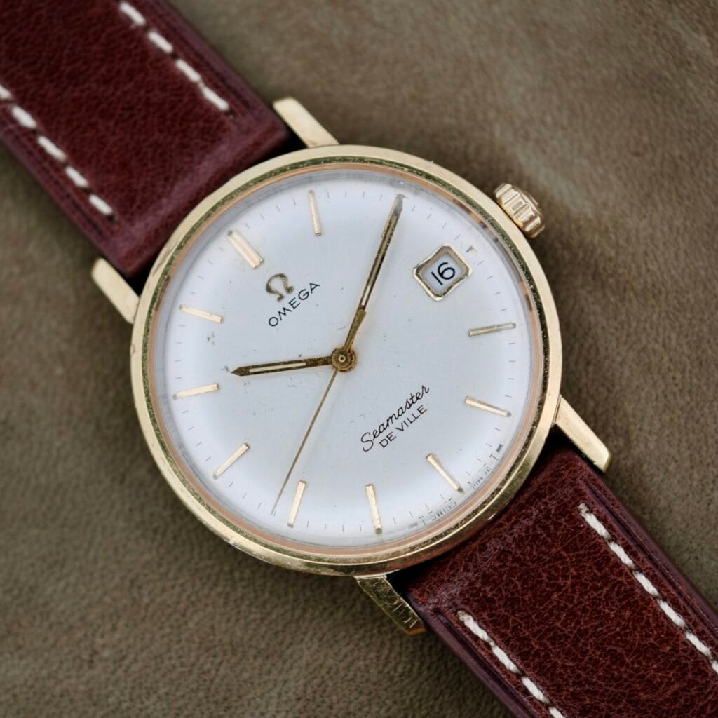 The Omega Seamaster Deville 155.020 Manual Wind is a vintage wristwatch featuring a gold case, white dial, date window at 3 oclock, and gold hour markers. It includes a brown leather strap with white stitching and is showcased on a beige surface.