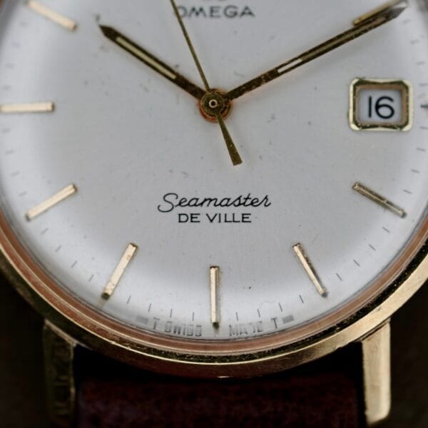 Close-up of an Omega Seamaster Deville 155.020 Manual Wind: a vintage watch with a white face, gold hour markers and hands, a date window displaying 16, encased in gold, and featuring a brown leather strap.