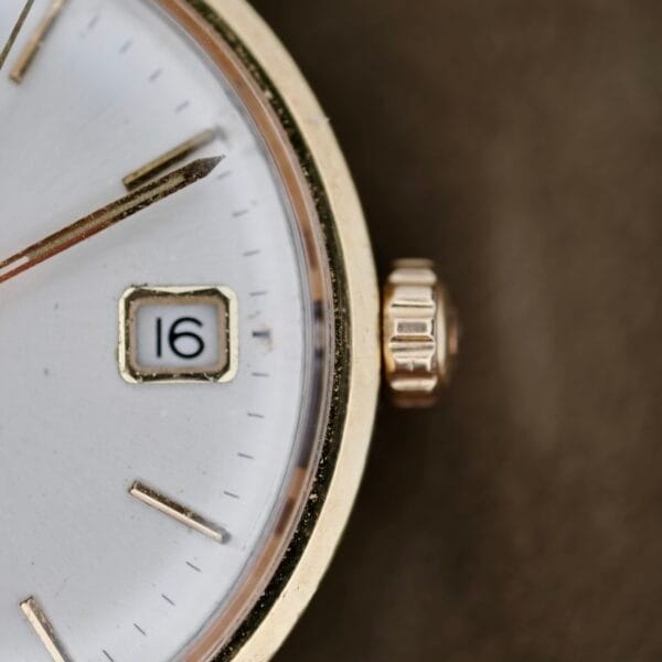 The close-up captures the Omega Seamaster Deville 155.020 Manual Winds gold wristwatch face, focusing on part of the dial and crown against a blurred background. The date window shows 16, with time hands partially visible on the left, highlighting its minimalist classic design.