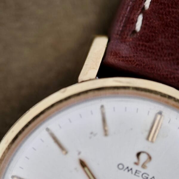 Close-up of an Omega Seamaster Deville 155.020 featuring a gold wristwatch with a white face, thin gold hour markers and hands, and the Omega logo. It has a brown leather strap with white stitching along the edge.
