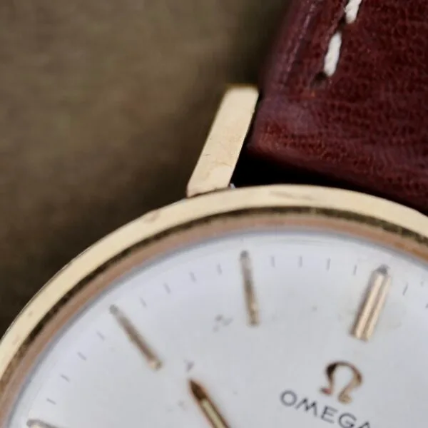 Close-up of an Omega Seamaster Deville 155.020 featuring a gold wristwatch with a white face, thin gold hour markers and hands, and the Omega logo. It has a brown leather strap with white stitching along the edge.