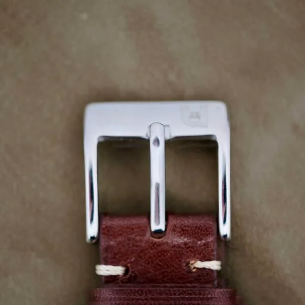 An image shows a shiny silver metal buckle attached to a brown leather belt, highlighted against a soft, light green background. White stitching secures the belt. Featured is the Omega Seamaster Deville 155.020 Manual Wind.