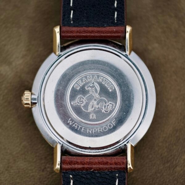 The image depicts the back of an Omega Seamaster Deville 155.020 Manual Wind wristwatch, featuring an engraved seahorse and Seamaster Waterproof inscription. Enhanced with a metal casing, it includes a black leather strap with brown stitching, set against a muted fabric background.