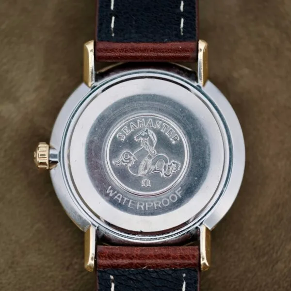 The image depicts the back of an Omega Seamaster Deville 155.020 Manual Wind wristwatch, featuring an engraved seahorse and Seamaster Waterproof inscription. Enhanced with a metal casing, it includes a black leather strap with brown stitching, set against a muted fabric background.