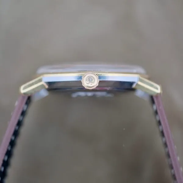 The side view of the Omega Seamaster Deville 155.020 Manual Wind displays its crown and case, featuring a two-tone gold and silver design. The strap looks like its crafted from leather, set against a blurred background to highlight the watch.