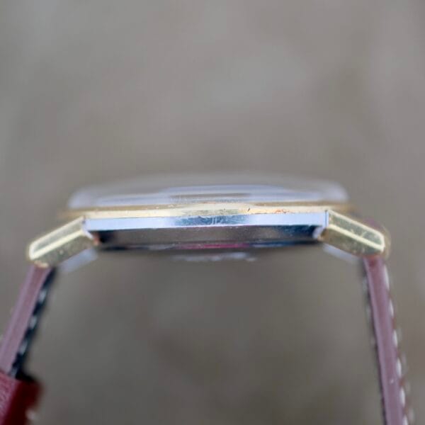 Side view of the Omega Seamaster Deville 155.020 Manual Wind. It features a metallic frame with gold-colored sides and housing, complemented by a partially visible brown leather strap attached to the case. The background presents a blurred, neutral color.