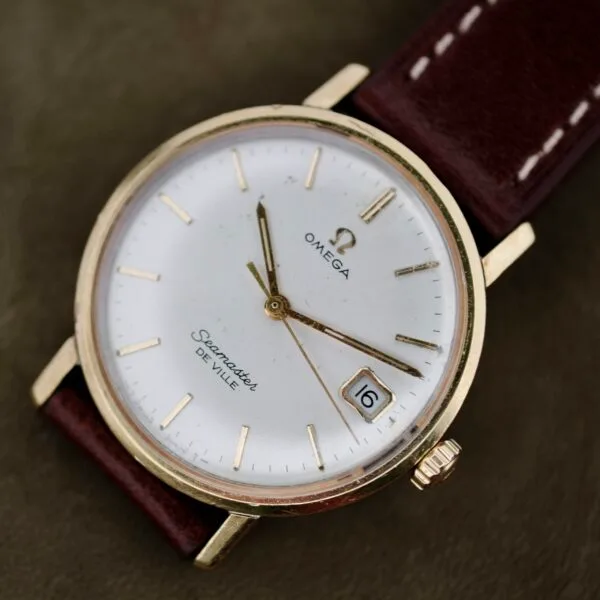An Omega Seamaster Deville 155.020 Manual Wind watch features a white face with gold hour markers and hands, a brown leather strap, and a date window showing the number 6. The crown is positioned on the right side.