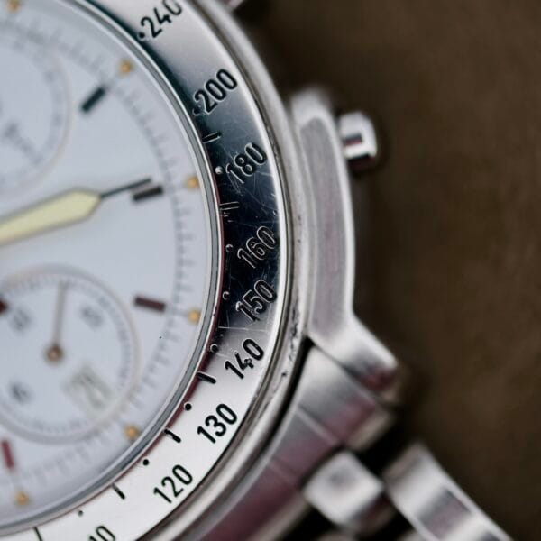 A close-up of the Baume Mercier Formula S Chronograph reveals a silver analog watch with a white dial, featuring a tachymeter scale on the bezel marked 100 to 240, three subdials, one central hand, and part of its metal bracelet.