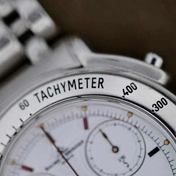 Close-up of the Baume Mercier Formula S Chronographs silver face, featuring a tachymeter scale, two subdials, and fine black markings. The word TACHYMETER is prominently displayed at the top, complemented by its metal bracelet.
