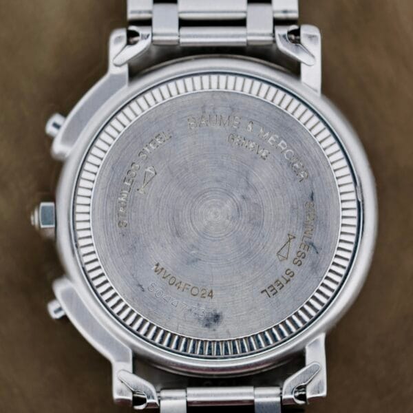 The image displays the back of a stainless steel Baume Mercier Formula S Chronograph watch. Engravings include BAUME & MERCIER, GENEVE, STAINLESS STEEL, and code MV04F024 5045455. It features a metal link band.