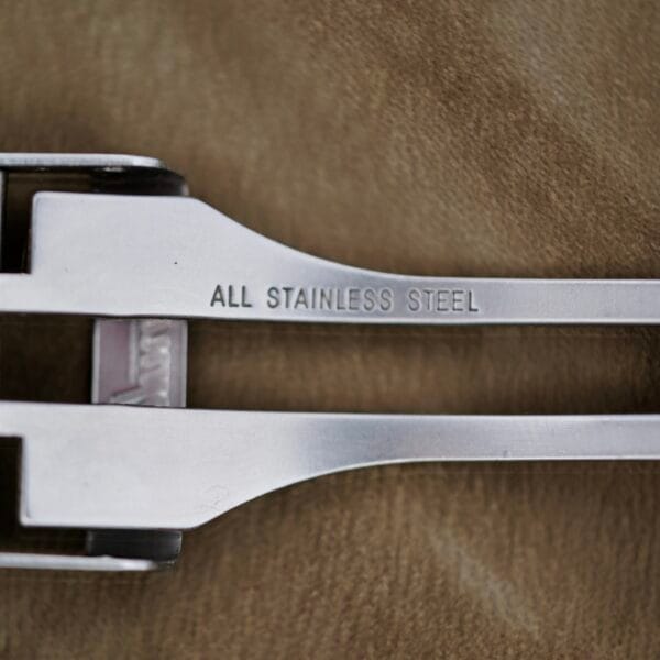 Close-up of two stainless steel utensils on a brown surface, with the top one engraved ALL STAINLESS STEEL, resembling the Baume Mercier Formula S Chronograph.