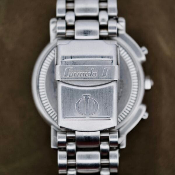 Close-up of the back of a metal wristwatch with a silver band. The clasp is engraved with Formula S and the Baume Mercier logo. The background is out of focus.