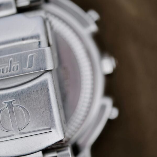 Close-up of the Baume Mercier Formula S Chronographs silver metal clasp with engraved initials and decorative elements, partially open to reveal a glimpse of the watch face. A blurred background enhances the watchs details.