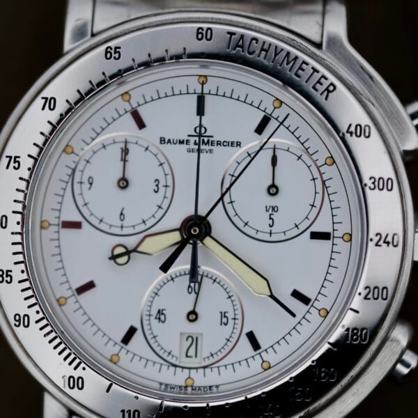 Close-up of a Baume Mercier Formula S Chronograph, silver with tachymeter bezel, white dial with three subdials, date window at 6 oclock, silver hour markers, and metal bracelet.