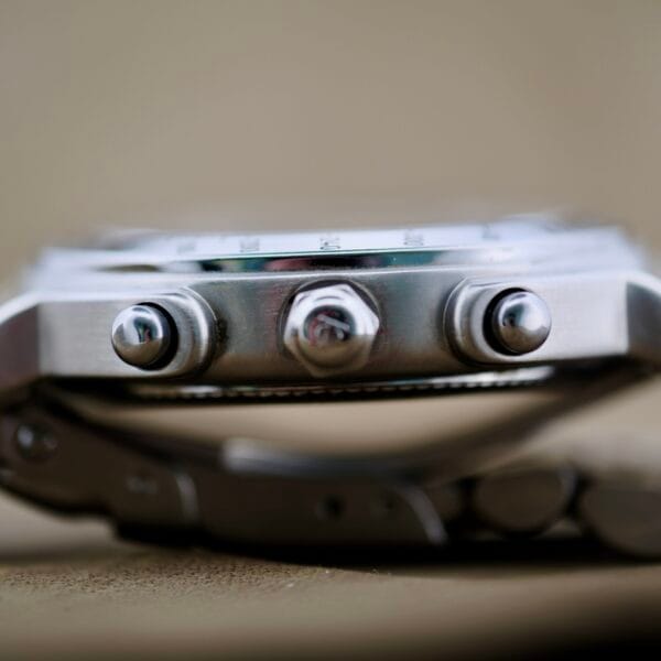 A close-up side view of the Baume Mercier Formula S Chronograph wristwatch showcases two round push buttons and a crown for time adjustment, featuring a brushed stainless steel finish against a blurred beige background.
