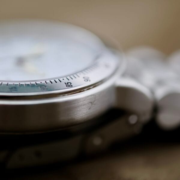 Close-up of the Baume Mercier Formula S Chronograph, showcasing the silver edge of its face and part of its bracelet. The bezel displays small, evenly spaced markings as the watch surface fades slightly out of focus to highlight the metals texture.