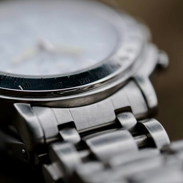 Close-up of the Baume Mercier Formula S Chronograph, highlighting the stainless steel metal link bracelet and part of the bezel with engraved numbers. The focus is on these details while the watch face is blurred, showcasing a muted silver tone.