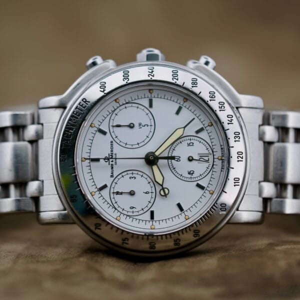 A close-up of the Baume Mercier Formula S Chronograph showcases its silver design with a tachymeter bezel, three sub-dials, and a date window. The blurred background emphasizes its detailed face and metallic band.