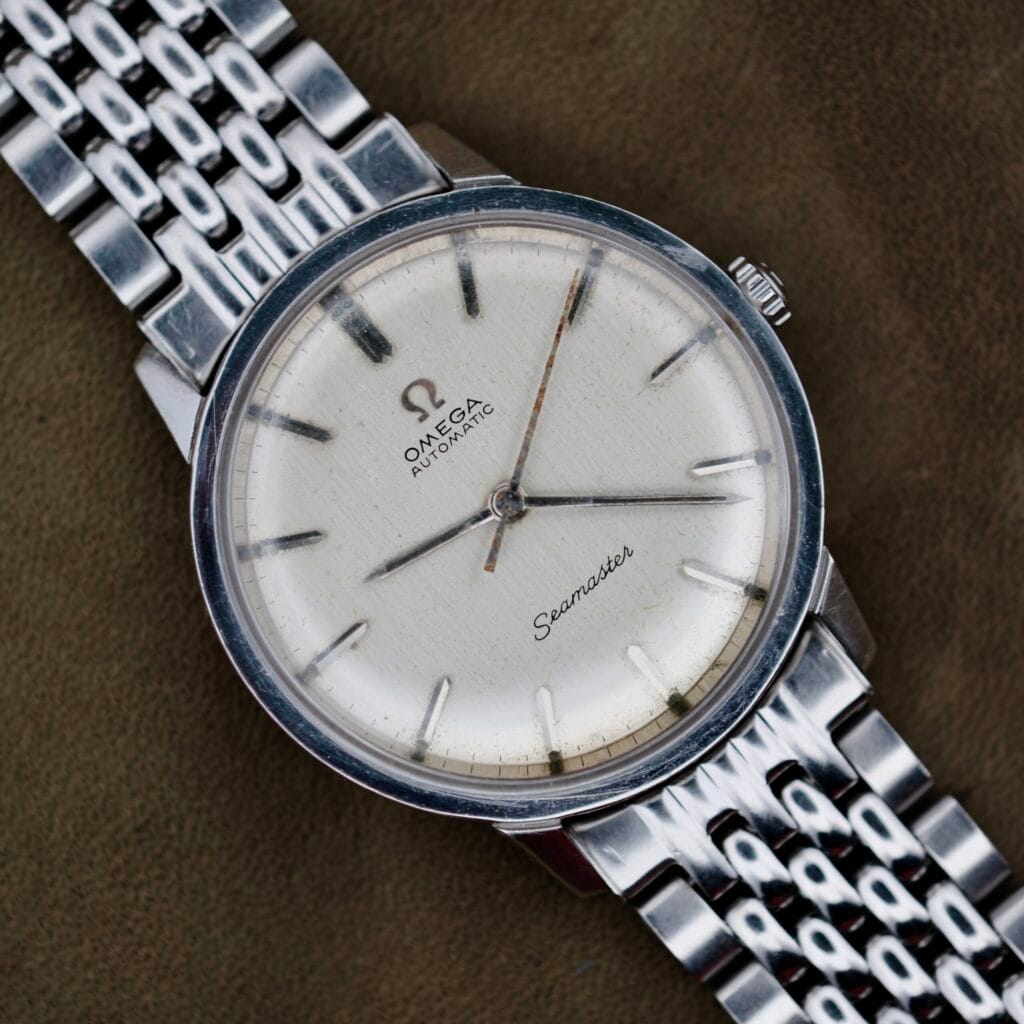 An Omega Automatic Seamaster wristwatch in silver features a round face with simple hour markers and hands, displaying the Omega logo and Seamaster name, set against a textured dark green fabric background. It comes with a metal link band.