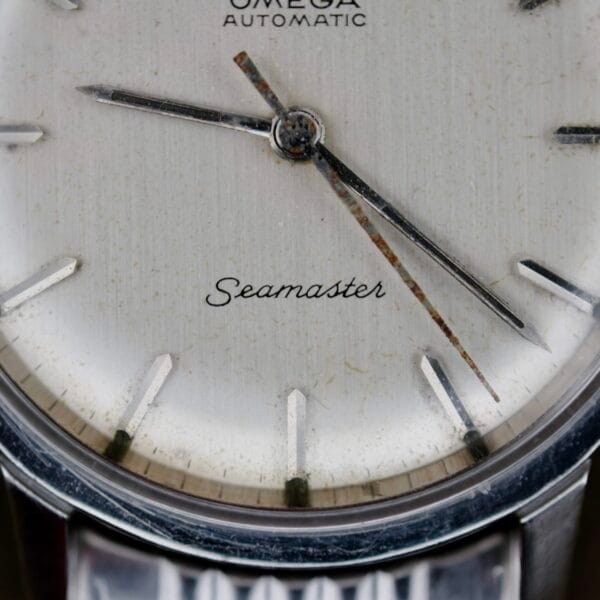 Close-up of the Omega Automatic Seamaster watch face, revealing a vintage patina. It displays a metallic dial with silver hour markers and slender hands, featuring Automatic below the logo and Seamaster in cursive.