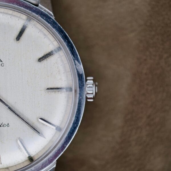 Close-up of an Omega Automatic Seamaster, featuring a vintage wristwatch face with a worn silver frame. It has a beige dial, black hour markers, minimalistic silver hands, and a visible winding crown on the right. The surface shows signs of wear and aging.