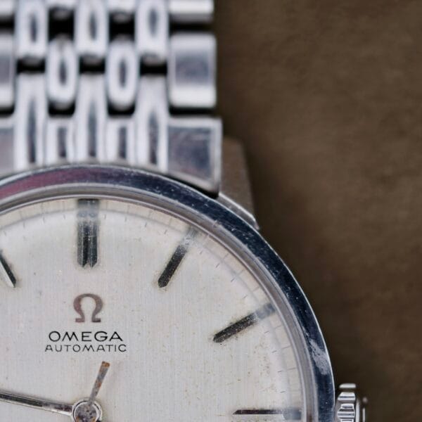 Close-up of a silver Omega Automatic Seamaster wristwatch featuring a stainless steel band. The off-white watch face displays minimalistic black hour markers and hands, with the Omega logo and AUTOMATIC clearly visible.