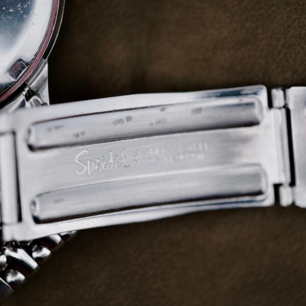 A close-up of a shiny metallic watch band engraved with Speidel, Stainless Steel, and USA. It features a folding clasp mechanism. The Omega Automatic Seamaster watch face can be partially seen at the images edge.