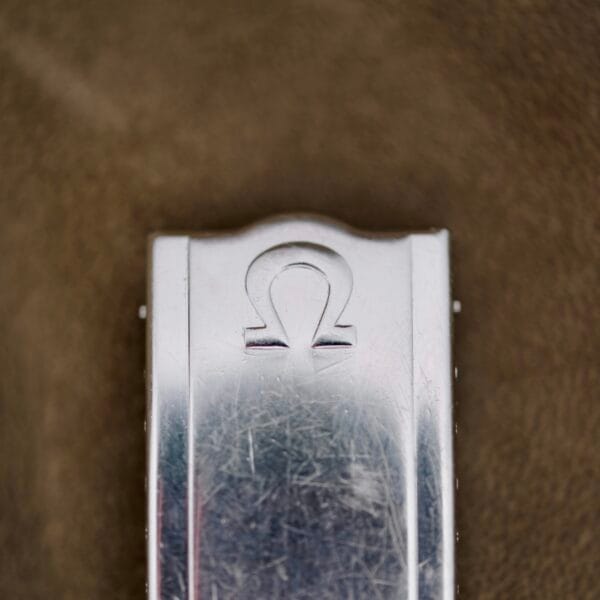 Close-up of a metallic clasp engraved with the Omega symbol on the Omega Automatic Seamaster, displaying visible scratches and a reflective quality against a textured, brownish background.