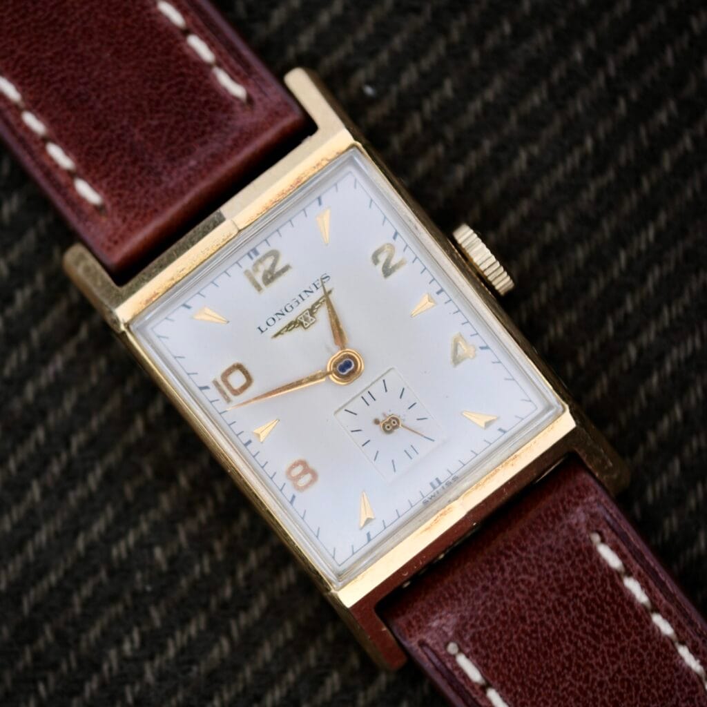 The Longines Tank Manual Wind 22L features a rectangular gold case, white dial, brown leather strap, and small seconds subdial, displayed on a dark fabric background.