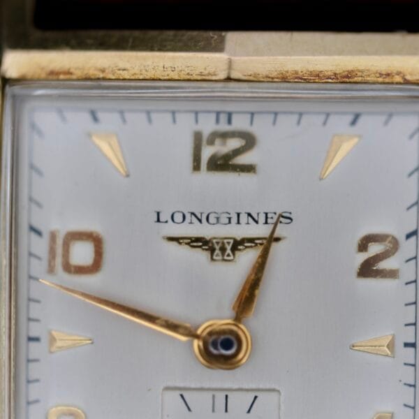 Close-up of the Longines Tank Manual Wind 22L watch face. It showcases gold hour indices and hands on a silver backdrop, with the logo below 12 oclock and a small sub-dial near 6 oclock, all encased in a luxurious gold case.