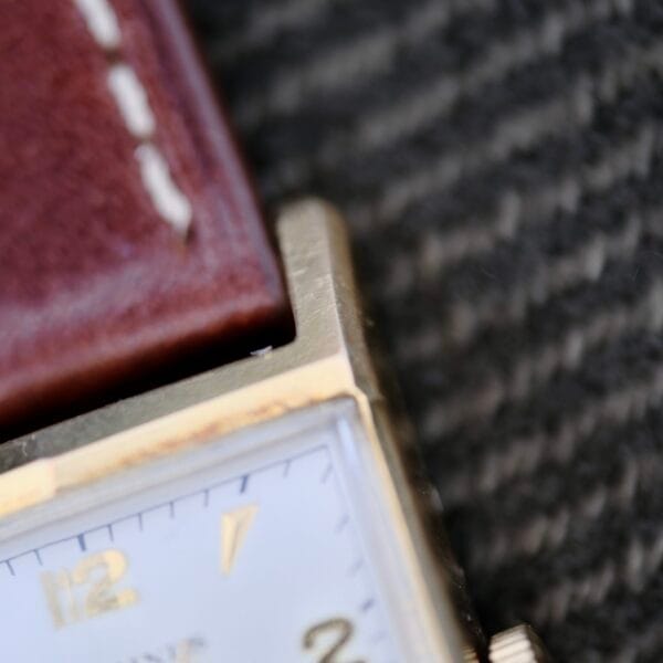 Close-up of a Longines Tank Manual Wind 22L watch featuring a gold rectangular casing and brown leather strap. The white dial with gold hour markers is elegantly set against the patterned fabric in the background.
