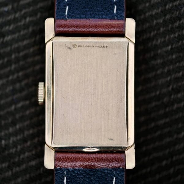 The Longines Tank Manual Wind 22L watch features a worn gold-toned rectangular case with 14K GOLD FILLED engraved on the back. It includes a brown leather strap with blue edges and white stitching, set against a textured dark fabric background that enhances its classic appeal.