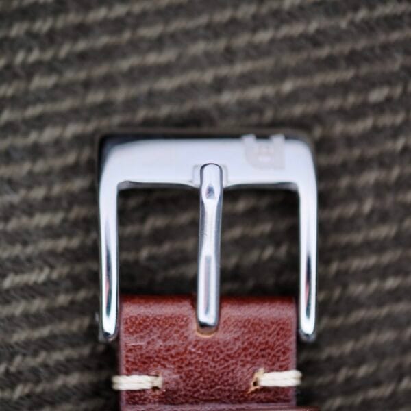 A shiny silver belt buckle, echoing the elegance of the Longines Tank Manual Wind 22L, is attached to a brown leather strap with beige stitching. The textured grayish backdrop enhances the compositions sophistication.