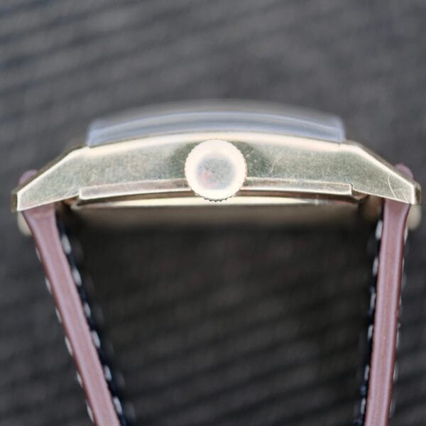 Close-up of a Longines Tank Manual Wind 22L, highlighting its rectangular metal case viewed from the side. It has a round crown and textured brown leather strap with visible stitching, set against a blurred dark background.