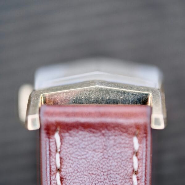 Close-up of a Longines Tank Manual Wind 22L, highlighting the metal case edge and a portion of the brown leather strap with visible stitching against a dark blurred background.
