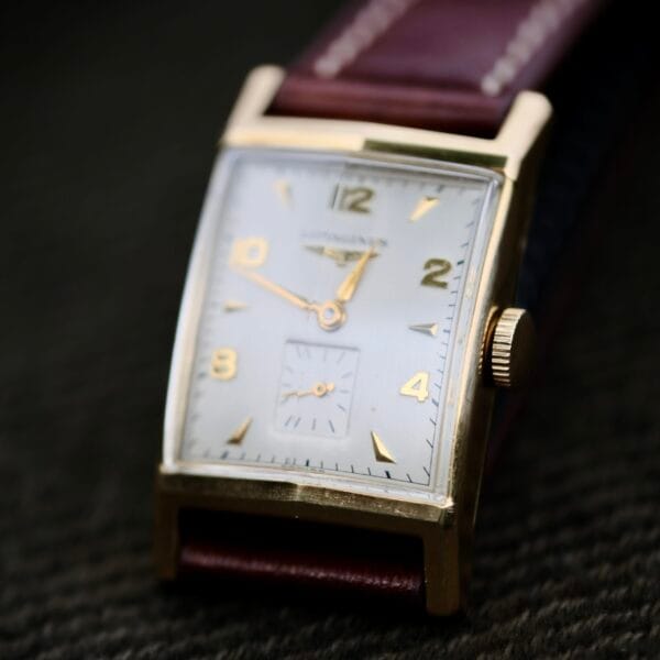 A close-up of the Longines Tank Manual Wind 22L wristwatch highlights its rectangular gold case and white dial, featuring gold numerals, hour and minute hands, a small seconds subdial, and an elegant dark leather strap.