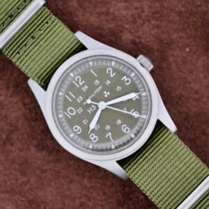 A Hamilton Khaki H3 Reference 9219 with a green nylon strap sits on a brown textured surface.