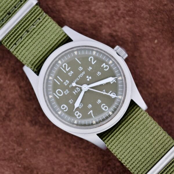 A Hamilton Khaki H3 Reference 9219 with a green nylon strap sits on a brown textured surface.